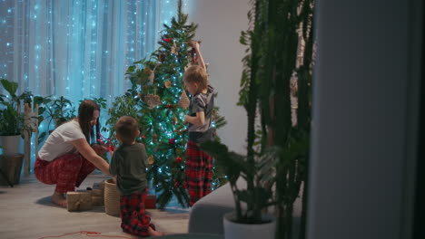 A-young-mother-and-two-boys-in-pajamas-decorate-the-Christmas-tree-with-toys-on-New-Year's-Eve.-Preparing-for-the-new-year-and-Christmas.-Decorating-the-house.-High-quality-4k-footage
